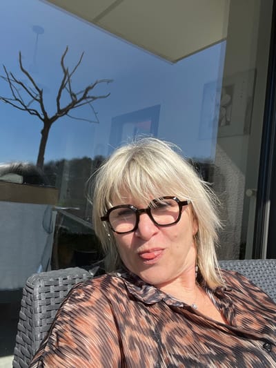A blonde-haired Caucasian female, wearing dark hexagonal glasses and a brown-and-black patterned blouse, is depicted seated outdoors in a dark grey woven chair under direct sunlight in Chambœuf, France. She is making a playful facial expression with her tongue slightly extended. Behind her, a large glass window reflects a bare-branched tree against a clear blue sky, distant foliage, and interior elements including a framed picture and a ceiling light fixture.