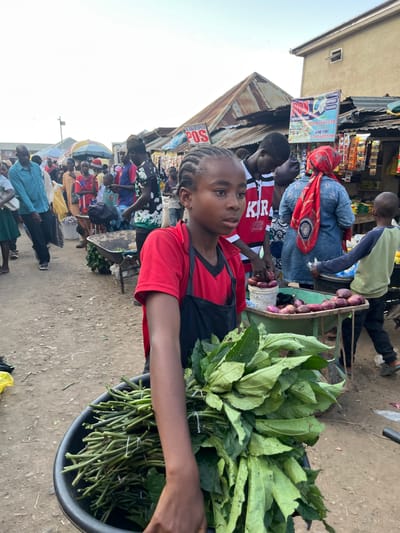 In Rafin Par, Nigeria, a busy market scene unfolds under the bright sky. The foreground is dominated by a young person wearing a red and black shirt, holding a large bunch of green vegetables in a black tub. Their gaze is directed towards the viewer, with a neutral expression. Behind them, a bustling marketplace is evident, with numerous people engaged in various activities.
Stalls are set up, displaying an array of goods, including what appear to be packaged snacks and other items on shelves. A woman in a blue top and a red headscarf is visible near one of the stalls, alongside other market-goers. Further back, more people are gathered, some appearing to be shoppers and others vendors. A child in a green shirt and dark pants walks past a stall laden with purple onions in a basket. The ground is dirt, with scattered debris, indicative of an outdoor market.
The overall impression is one of a lively, everyday market day. The bright natural light suggests it is daytime, likely mid-morning or afternoon. The presence of diverse people and the arrangement of goods point to a commercial hub where daily necessities are bought and sold. The setting appears to be an open-air market, common in many Nigerian towns and cities. The emotional tone is neutral, focused on the candid capture of a moment in a vibrant community. Visible text includes "POS" on a sign in the background, indicating a point-of-sale service, and numbers on a sign which may relate to contact information for a business.