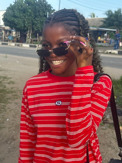 A young Black woman, with braided hair and long pink fingernails, is shown from the chest up, smiling and looking at the camera. She is wearing black sunglasses and a red and white striped long-sleeved shirt with a small oval logo on the left chest. She has rings on her fingers, one of which is ornate with colorful stones. The background appears to be a street scene in Badagry, Nigeria, with a road, some buildings that look like shops or stalls, and trees. The lighting suggests it is daytime.