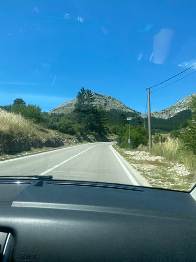 The image depicts a road scene taken from the interior of a vehicle, likely in Kopito, Montenegro. The foreground shows a portion of the car's dashboard, including the textured plastic and a visible "SRS AIRBAG" label. The windshield reveals a clear blue sky with some reflections, and a pair of windshield wipers.
The road ahead curves to the left, with a white line marking the center and a white edge line. The roadside features dry, golden grass on the left and more lush green vegetation, rocks, and trees on the right. In the middle ground, a wooden utility pole with wires is visible, along with a small solar-powered device mounted on a pole. Beyond the road, hills covered in dense green forest rise, with a rocky peak dominating the background. The overall environment suggests a rural or mountainous area under bright daylight.