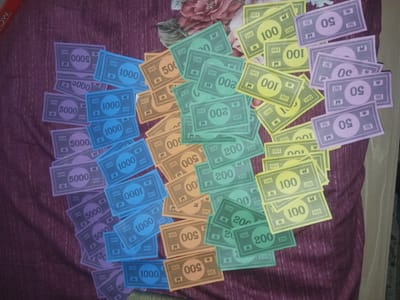 The image displays various denominations of colorful Monopoly game money scattered across a dark purple fabric surface. This surface features a subtle striped texture and a floral pattern visible towards the top right.
The Monopoly money is generally grouped by color and denomination, though arranged somewhat haphazardly. Visible denominations include:
- Approximately 10 purple 5000 notes.
- Around 12 blue 1000 notes.
- Roughly 10 orange 500 notes.
- About 15 green 200 notes.
- Approximately 14 yellow 100 notes.
- Around 6 pink/light purple 50 notes.
Each note prominently displays its numerical value, often with smaller repeating numbers and the characteristic Monopoly "H" symbol. Some bills are partially obscured or oriented upside down. In the top-left corner, a red object, likely a game box, is partially visible with the text "MO", hinting at the "Monopoly" brand. The scene, located in Gwarinpa, Nigeria, appears to be indoors with ambient lighting, suggesting the money is laid out either before or after a board game session.