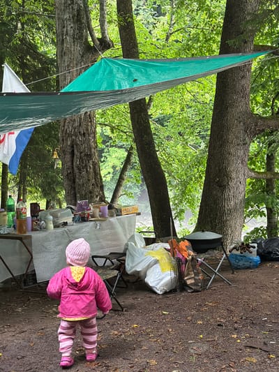 A child, viewed from behind, wearing a pink knit hat, a bright pink hooded jacket, and striped pink and white pants, stands on a dirt path in a forested area. A table laden with various items, including bottles of liquid, containers, and dishes, is set up in the background. Large tarps are stretched between trees, providing shade over the area. Several large bags and a black pot are also visible on the ground. The environment suggests a campsite or outdoor gathering.