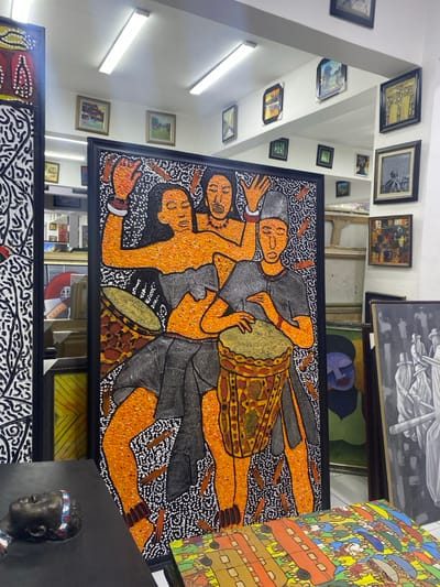 This image depicts an interior view of an art gallery or studio located in Alaguntan, Nigeria. The central focus is a large, vertical framed painting rendered in a textured, pointillistic style, featuring three stylized figures against a black background. The figures, depicted with orange-brown skin tones and black outlines, include two female figures with raised arms, one wearing a necklace, and a male figure in a grey tunic and cap actively playing a patterned drum. A second drum is partially visible to the left of the male figure.
The gallery space features white walls and ceiling illuminated by multiple fluorescent strip lights. Numerous framed artworks of varying sizes and styles, including landscapes and abstract pieces, are displayed on the walls. In the foreground, to the left, a portion of another large vertical painting with black and white abstract patterns is visible. A dark-colored bust or sculpture of a head, marked with red, white, and blue stripes on its forehead, rests on a dark surface in the immediate foreground. To the right, several paintings are stacked or leaning, including a grayscale work depicting multiple figures, a stylized human face in blue and green hues, and a colorful abstract composition. The floor is light-colored.
