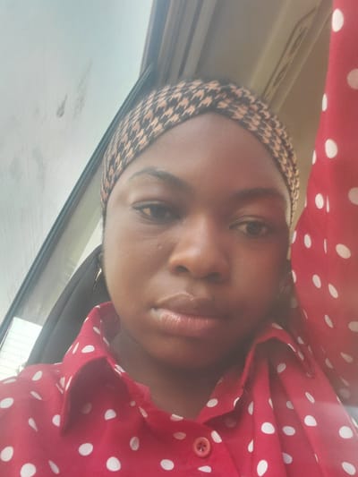A close-up, medium shot shows a young Black woman from the chest up, facing forward and slightly to the left. She is wearing a red, polka-dotted button-down shirt and a patterned headscarf. The background is blurred and appears to be the interior of a vehicle, with a window on the left side showing a faint reflection of what might be a sky. The lighting is soft and diffused. The woman's facial expression is neutral.