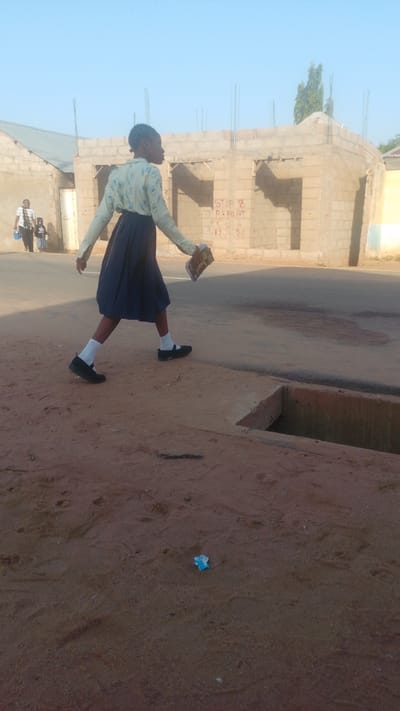 A young girl in a light blue patterned shirt and dark blue skirt walks on a dirt path in front of a building under construction in Tempure, Nigeria. She is wearing white socks and black shoes and is holding a book. The building is made of concrete blocks and has rebar sticking out of the top. In the background, another person and two children are walking away from the camera. The sky is clear and blue. There is a drainage ditch in the foreground. The ground is covered in sand and some small debris.