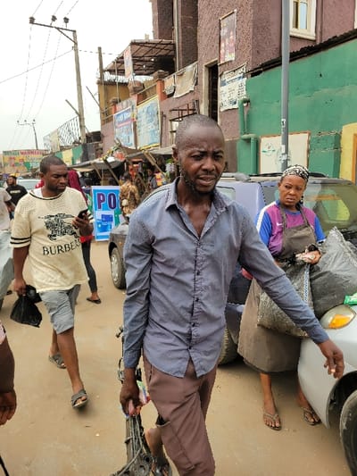 The image displays a bustling street scene in Onitsha, Nigeria, characterized by pedestrian traffic and commercial activity under an overcast sky. In the foreground, an adult male with a short, dark beard and shaven head walks towards the viewer, his gaze directed forward. He wears a dark blue-grey long-sleeved shirt, unbuttoned at the collar, and brown trousers, holding a metal object, possibly a mechanical component, in his right hand. To his immediate left, another adult male walks away from the viewer, wearing a cream-colored t-shirt with "BURBERRY" text, light grey shorts, and dark sandals. He is looking down at a smartphone in his left hand while carrying a black plastic bag in his right. To the right of the central figure, an adult female with a patterned headscarf, purple t-shirt, and dark brown apron stands beside a silver sedan, attending to items near the vehicle, including a large grey bag.
The background features multiple multi-story buildings of varying colors, including shades of brown, green, and yellow, lining the street. Numerous commercial signs are affixed to these structures, one prominent yellow sign on the far left reads "OGENE MAJMAJMA'S SUPER MARKET." Utility poles with overhead power lines are visible on the left, and a grey utility pole stands on the right. The ground is an unpaved or dirt-colored road surface. Several other individuals are partially visible in the midground and background, indicating a populated area. Vehicles, including a dark-colored car, are parked or moving within the scene.