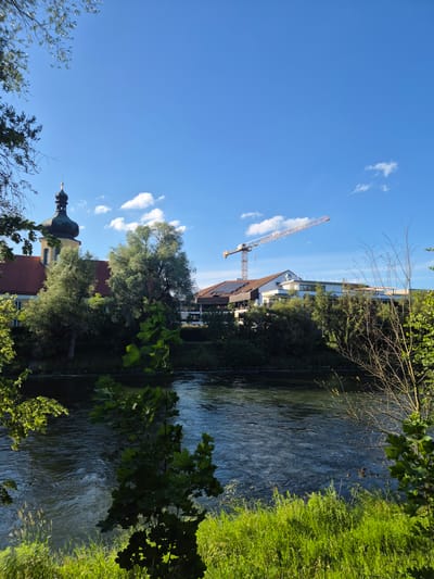 The image depicts a scenic view of the Isar river in Landshut, Germany. On the left bank, a building with a distinctive onion-domed tower, likely a church or monastery, is partially visible behind lush green trees. Across the river, on the right bank, a modern building with a white facade stands prominently. A tall construction crane is positioned over this building, suggesting ongoing development. The river itself is calm with gentle ripples reflecting the bright blue sky dotted with white clouds. The foreground is filled with vibrant green grass and foliage, framing the entire scene. The sun appears to be shining, casting shadows and illuminating the landscape.