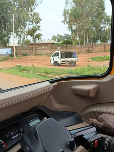 The image, captured from the interior of a three-wheeled vehicle, shows a street scene in Barakin Angundi, Nigeria. The foreground is dominated by the vehicle's dashboard and a hand resting on the steering mechanism. Through the side window, a dirt road is visible, with a white truck carrying several large drums in the middle ground. A low brick wall runs along the side of the road, with some graffiti on it. Beyond the wall, trees and buildings suggest a semi-urban or rural setting. The sky is overcast.