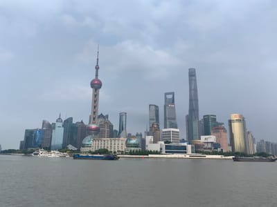 This media file captures a panoramic view of the Shanghai skyline, specifically the Pudong district, as seen from the Huangpu River. The scene is dominated by a collection of modern skyscrapers of varying heights and architectural styles, showcasing a bustling urban landscape.
Prominent among the structures is the Oriental Pearl TV Tower, an iconic landmark with its distinctive spherical design and slender spire. Adjacent to it are other notable buildings like the Shanghai Tower, a supertall skyscraper, and the Jin Mao Tower, as well as the Shanghai World Financial Center with its characteristic trapezoidal opening. The architecture is a blend of glass, steel, and concrete, with many buildings featuring sleek, contemporary designs.
The river in the foreground is calm, with several vessels visible. Two white passenger ferries are docked or cruising near the shore, and a larger blue barge is moving along the waterway. The water itself is a muted gray, reflecting the overcast sky.
The weather appears to be cloudy or overcast, with a soft, diffused light, suggesting it might be early morning or late afternoon. The sky is filled with layers of gray clouds, creating a somewhat subdued atmosphere.
The overall impression is one of a dynamic and impressive city, a testament to modern development and economic power. There is no visible text of significance in the image.