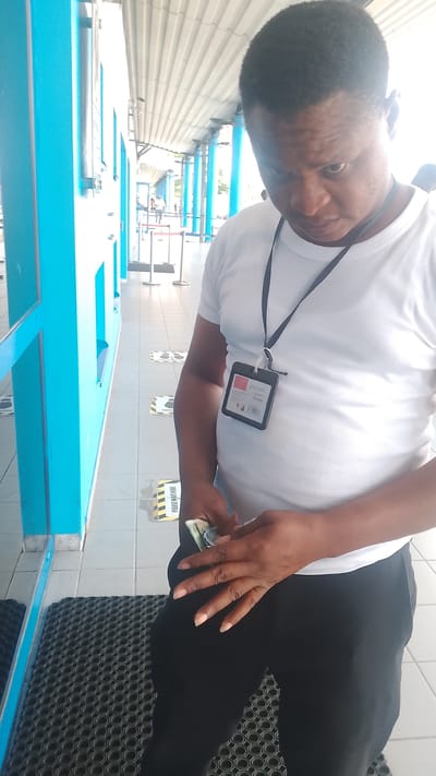 The media file shows a man in Orerokpe, Nigeria, wearing a white t-shirt and black pants. He has an identification card hanging from a lanyard around his neck, and is holding some cash in his hands. The scene appears to be an outdoor walkway with a bright, overcast sky, suggesting it might be daytime with diffused light.
The background shows a bright blue wall on the left, and a covered walkway with a corrugated metal roof supported by blue pillars. The ground is tiled, and there are social distancing markers on the floor with the text "PLEASE WAIT HERE". In the distance, other people can be seen walking in the walkway, and there are some red stanchions, possibly indicating a queue or a restricted area.
The man is looking down at the cash he is holding, and his expression is focused. The overall setting suggests a public area, possibly an entrance to a building or a service area where queuing and identification might be required. The presence of the cash and the identification card suggests an activity involving a transaction or an employee performing a duty. The weather appears to be fair. The text on the ID card is partially visible and seems to say "IDC CARD" and "No 8125".