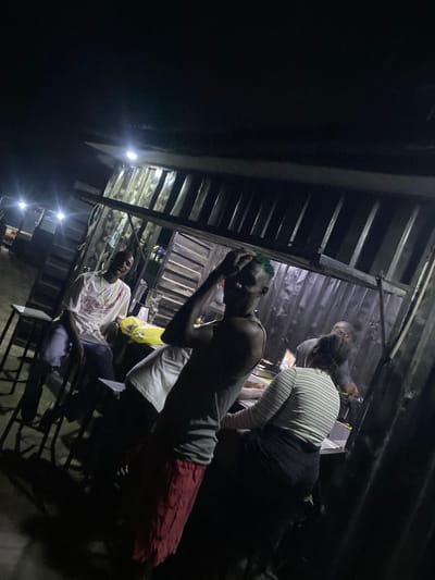 The image depicts a nighttime scene of several individuals gathered under an informal, semi-outdoor structure in Ado, Nigeria. The setting appears to be a shed or stall with a corrugated metal roof and exposed metal beams.

In the foreground, a young man with distinctive green-dyed hair is seated or leaning, wearing a dark tank top and red shorts. He holds his head in his hands, suggesting a pensive or possibly distressed emotional state. Behind him, another person in a white and black striped top and dark bottoms is seen leaning forward. Further back and to the right, a man in a white t-shirt and light-colored trousers is seated, looking towards the viewer. Other figures are dimly visible in the background, some interacting or standing.

The primary illumination comes from several bare, bright light bulbs, which cast harsh light and deep shadows across the scene, indicating it is nighttime. The artificial lighting creates strong glare in parts of the image. Visible objects include simple chairs or stools on which people are seated, and various blurry items in the background, including what looks like a yellow plastic bag. One person appears to be holding a glass.

The overall atmosphere suggests a casual social gathering or waiting area. The architectural style and informal setup are consistent with roadside establishments or community spaces in the region. No discernible text is visible within the frame. The photograph is slightly tilted, contributing to a candid feel.