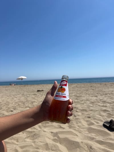 A person holds a bottle of orange-flavored basil seed drink on a sandy beach in Martil, Morocco, under a clear blue sky. The Mediterranean Sea stretches horizontally in the background, meeting the beach. A white beach umbrella and a few people are visible in the distance near the water. A black shoe rests on the sand. The drink bottle is transparent, revealing the basil seeds within the orange liquid. The label indicates it is "Basil Seed Drink with Orange Flavor."