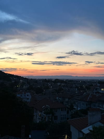 The image depicts a twilight view of the city of Asenovgrad, Bulgaria. The sky is filled with dramatic clouds, transitioning from deep blue at the top to vibrant oranges and pinks near the horizon, indicating sunset or sunrise. Below the sky, the cityscape is visible, characterized by a dense arrangement of buildings with red-tiled roofs. The buildings appear to be primarily residential, with varying architectural styles, and some show illuminated windows. The terrain slopes upwards towards the left side of the frame, where a forested hill is prominent, casting shadows over the buildings at its base. The overall atmosphere is tranquil and serene, capturing the quiet beauty of the city as day transitions to night. There are no people or discernible actions captured in the scene.