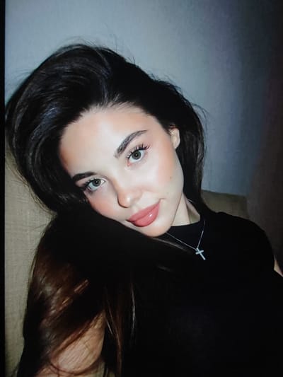 The image shows a close-up, head-and-shoulders portrait of a young woman with dark hair, fair skin, and green eyes. She is wearing a black top and a silver cross necklace. Her head is tilted, and her hair is falling around her face. The background is dimly lit and appears to be an indoor setting. There is no information in the image or provided context that would allow for its location to be identified within Fergana City, Uzbekistan, or any other specific geographic location.