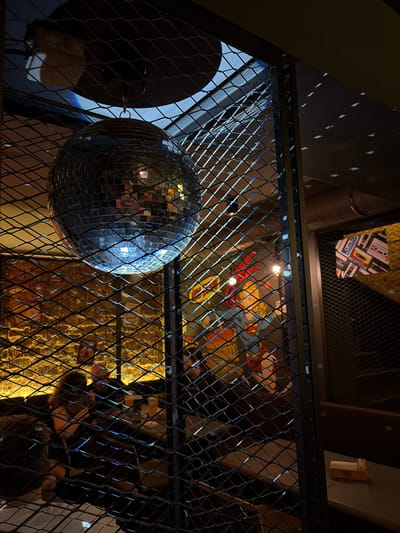 An indoor, dimly lit scene features a disco ball suspended by a chain, reflecting light in a room with a brick wall and a metal mesh partition. Through the mesh, several people are visible in what appears to be a seating area, possibly a bar or restaurant. A decorative item depicting cassette tapes hangs on the wall in the background, and a small, yellow dispenser is present on a surface to the right. The overall atmosphere suggests a casual, social setting. The location is specified as Minsk, Belarus.