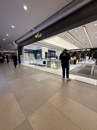 This media file captures an indoor scene within a brightly lit, modern retail environment, likely a shopping mall in Bangkok, Thailand. The primary subjects are several individuals shopping or working in an electronics store. One person in the foreground, seen from behind, wears a dark green t-shirt with text that reads "Make Your Moment." Another person, dressed in black, is also visible in the lower part of the frame.
The scene is set in a store that appears to sell mobile phones and accessories, indicated by prominent signage for "AIS," "SAMSUNG," "UGREEN," and "OPPO." Display shelves filled with products are visible, as are illuminated screens showing advertisements. The flooring is tiled in a neutral, light-colored stone. The lighting is bright and artificial, suggesting it is either evening or the interior is designed to be well-lit regardless of the time of day. The overall atmosphere seems busy and commercial, typical of a retail setting.