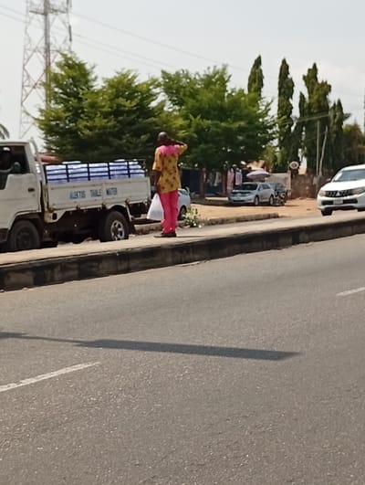 The image depicts a daytime street scene in Adankolo, Nigeria. A two-lane asphalt road occupies the foreground, featuring a dashed white line on the right side and a long shadow extending across it. A dark gray concrete curb separates the road from a dirt shoulder.
On the dirt shoulder, a white light-duty truck is visible on the left, parked or moving slowly. Its open bed is loaded with multiple rows of packaged bottled water, likely in blue or white wrapping. The side of the truck bed bears the inscription "ALEKTUS TABLE WATER." A driver's side cabin is partially visible.
Standing to the right of the truck on the dirt shoulder is a bald male subject with dark skin. He wears a short-sleeved shirt patterned in yellow and pink, and matching pink trousers. His right hand is raised to the back of his neck or head, and he holds a white plastic bag in his left hand. He faces away from the viewer, looking towards the right side of the frame.
Across the dirt shoulder, a commercial area is visible. It features small structures or stalls with items, possibly clothing, hanging for display. A silver sedan, another dark vehicle, and a motorcycle are parked on a dirt lot behind these structures. A red circular sign is also discernible. A white SUV is partially visible on the main road in the far right foreground.
The background consists of green trees, including large, leafy trees on the left and taller, slender trees on the right. A large metallic lattice tower, indicative of a high-tension power line, is prominent in the upper left background. The sky is bright and clear.