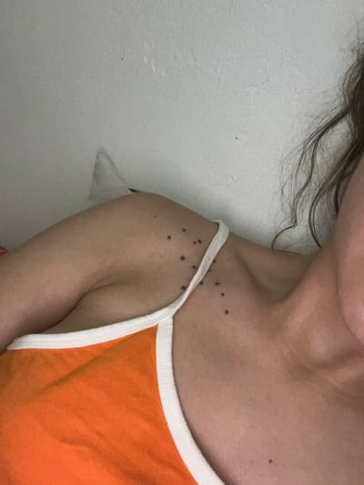 A close-up photograph shows a partial view of a person's upper body. The person has fair skin and brown hair. The person is wearing an orange tank top with white trim. There are several small, black star-shaped tattoos on the person's shoulder. A mole is visible on the person's chest and shoulder. The background is a white wall. The image does not contain sufficient information to determine a specific location in Saarbrücken, Germany.