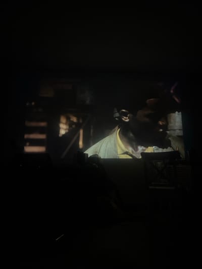 The image depicts a dimly lit interior in Pedrouços, Portugal, with a projected video occupying the upper half of the frame. The projected content shows a dark-skinned individual, likely male, from the chest up, facing right. He wears a light-colored top, possibly yellow or beige, and has short, dark hair; his mouth is slightly open. The background of the projection is dark and features architectural elements, including a vertical, ladder-like structure on the left and a warm light source or opening in the center. The lower half of the room is in deep shadow, revealing faint silhouettes of domestic objects. On the far right, a discernible object resembling an hourglass or a multi-tiered stand is present on a surface. A dark, indistinct mass in the lower-center suggests a seated figure or furniture. The only significant light source emanates from the screen projection, casting the rest of the room into profound darkness. No explicit actions or interactions are clearly discernible within the immediate viewing environment.