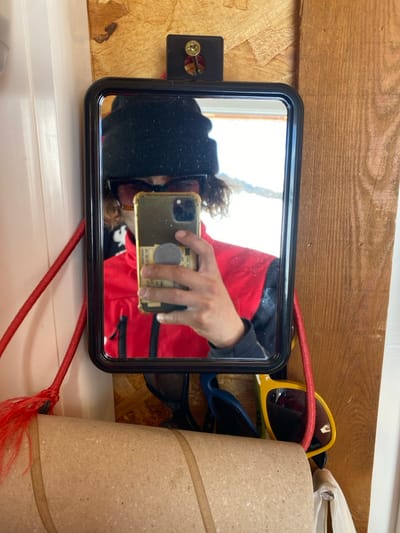 The image displays a mirror selfie taken by an individual in an indoor setting in La Salle-les-Alpes, France. A black-framed, rectangular mirror with rounded corners is mounted on a light-colored oriented strand board (OSB) wall, secured by a black bracket and a gold-colored screw at the top. The individual, wearing a black beanie, dark sunglasses, and a red jacket over a black garment, is reflected in the mirror. Their right hand holds a gold-cased smartphone with a grey pop socket, capturing the reflection. A portion of a banknote is visible tucked behind the pop socket. Curly dark hair extends from beneath the beanie. The mirror's reflection reveals a bright, snow-covered outdoor landscape, indicative of an alpine environment. To the left of the mirror, a braided red cord with a fringed end hangs downwards. Below the mirror, several objects are visible: at least two pairs of sunglasses, one dark and one bright yellow with a red elastic strap, and a large, horizontal cardboard tube in the immediate foreground. A vertical plank of darker wood is visible to the right of the mirror, and a crumpled white plastic bag is partially visible in the lower right corner.