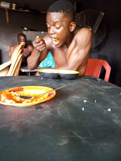 The image depicts a scene inside a dimly lit room, likely in Onitsha, Nigeria. In the foreground, a young, shirtless man with dark skin and short hair is actively eating from a black bowl, holding food in his right hand and bringing it towards his mouth. His eyes are closed as he chews. In the background, another shirtless man with dark skin and a shaved head is partially visible, seated and looking down. To the left of the foreground man, a woven basket with yellow-brown material spills over. A red chair is visible behind the foreground man, and a large, dark fan is mounted on the wall behind that. The foreground is dominated by a dark, textured table with a splatter of white grains scattered across its surface. An orange plate with remnants of food and a spoon rests on the table, closer to the viewer. The overall environment appears utilitarian and somewhat sparse.
