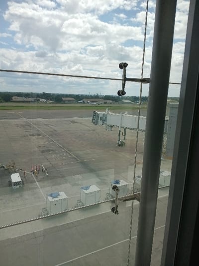 This image depicts a view from within an airport terminal at Robert Gabriel Mugabe International Airport in Harare, Zimbabwe, looking out onto the apron and distant runway under a cloudy sky. The foreground is partially obscured by structural elements of the terminal's glass window, including a vertical grey metal pillar on the right and a horizontal tension cable secured by four-bolt glass clamps across the middle.

On the grey asphalt apron, a white, stationary jet bridge is visible, positioned without an aircraft attached. To the left of the jet bridge, ground support equipment includes a white baggage cart with red railings and a cluster of red and white traffic cones and associated equipment. Closer to the viewer and below the jet bridge, three white cargo containers, each topped with a blue cover or tarp, are arranged in a line on ground dollies. Further in the background, a green strip of grass separates the apron from a distant runway or taxiway where parts of a white aircraft and low-rise buildings are discernible. The horizon features a distant treeline, scattered buildings, and a tall, slender mast or tower. No individuals or explicit actions are observed within the frame.