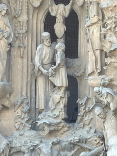 This is a close-up, vertically oriented, desaturated photograph of a bas-relief sculpture. The sculpture appears to be made of stone and depicts a scene with multiple figures. The figures are carved in a detailed, somewhat stylized manner, with flowing robes and expressive faces. The style suggests it is religious or historical in nature, potentially depicting a narrative from a biblical story or a historical event.

The central focus of the image is on two figures lying down, seemingly deceased or in repose, with other figures surrounding them. One of the prominent figures on the left is wearing a headdress and appears to be dressed in ceremonial robes. Another figure is being held or supported by someone above. The overall scene conveys a sense of solemnity or grief, with the figures appearing to be part of a burial or a significant event of mourning.

The background behind the figures is a dark, gridded surface, possibly indicating a window, grate, or an opening in the architectural structure the sculpture is part of. The lighting is diffuse, casting soft shadows and highlighting the textures of the stone. There are no discernible text elements within the frame. The sculpture's intricate detail and the subject matter point to a significant artistic and cultural work, likely part of an architectural facade. Given the location context of Barcelona, Spain, this could be a detail from a renowned Gaudi building or a similar gothic or modernist architectural masterpiece.