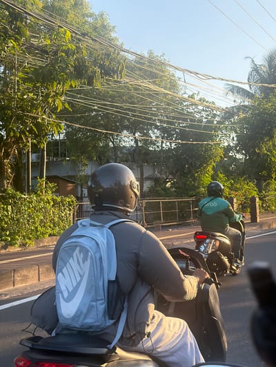 The image shows two people riding motorcycles on a street in Kuta Selatan, Indonesia. The foreground shows the back of a person wearing a helmet and a light blue backpack with a Nike logo. They are riding a dark-colored motorcycle. Behind them, another person is riding a motorcycle and wearing a green jacket with a "Grob" logo on the back. The background features trees, power lines, and buildings. The lighting suggests it is either early morning or late afternoon, with sunlight filtering through the trees. The road appears to be paved, and there is a sidewalk with a low concrete barrier.