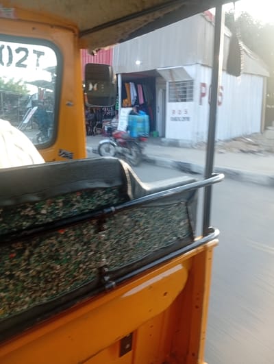 The image was taken from inside a yellow auto-rickshaw in Jalingo, Nigeria. The auto-rickshaw has a black seat with a patterned underlayer. Through the windshield, a white building with a sign that reads "POS" and "CHOCONIC" is visible. Next to the building are racks of clothing. A motorcycle is parked in front of the building. The road and sidewalk are visible in the background. The sun is shining, casting a bright glare on the scene.