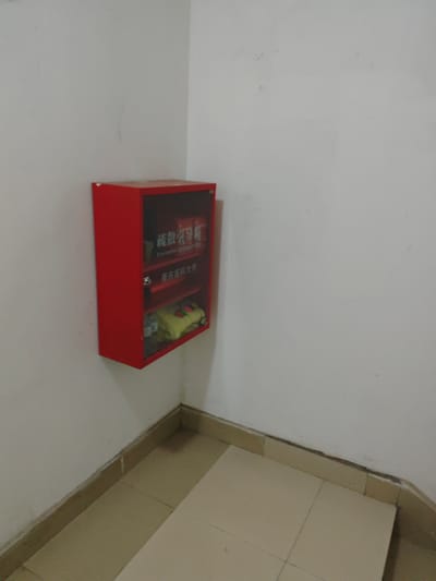 A red, wall-mounted cabinet with a glass front is located in a corner of a room with white walls and beige tiled flooring. The cabinet, identified by Chinese characters on its front as an "Evacuation Emergency Box" associated with "Chongqing Medical University," contains yellow fabric items. The scene appears to be an interior hallway or common area within the Chongqing High-tech Zone, China.