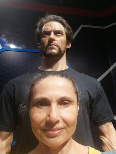 A photograph depicts a woman posing closely in front of a wax figure of a male, taken in the city of Hong Gai Ward, Vietnam. The woman, who is positioned in the foreground, is smiling slightly. The wax figure of the man, positioned behind the woman, has dark hair and a beard. The wax figure wears a black t-shirt. The background features a dark blue and black wall with a hexagonal pattern.