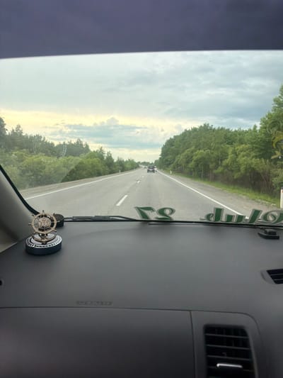 The image is taken from the perspective of a person inside a vehicle, looking out through the windshield. The vehicle appears to be on a paved, two-lane road with a white line marking the center and the right edge. Trees line both sides of the road, with a dense forest on the right and sparser trees on the left. The sky is cloudy with patches of yellow and white, suggesting either sunrise or sunset.
On the dashboard of the vehicle, in the foreground, there is a decorative item that resembles a ship's steering wheel mounted on a circular base. To the right of this item, there are green decorative letters applied to the dashboard.
Further down the road, several cars can be seen in the distance, indicating traffic. The environment appears to be rural or on the outskirts of a town. The weather seems overcast.
The location is identified as the city of Приамурский, Russia.