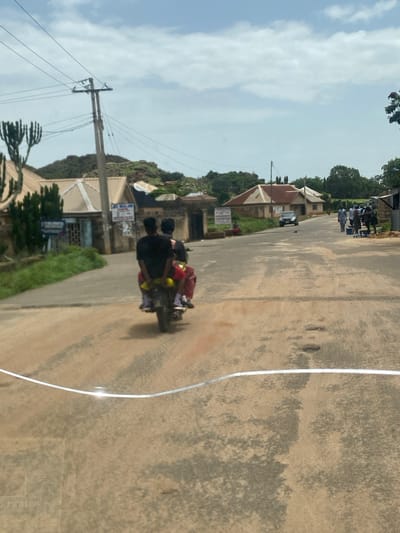 A motorcycle carrying three people travels along a paved road in Bukuru, Nigeria. The rider wears a black shirt and a red backpack. The two passengers in the back also appear to be wearing dark clothing. The motorcycle has a yellow object on the back, possibly a helmet or bag, and the passengers are wearing checkered shoes.
The scene appears to be a residential area with houses lining the road. Some houses have corrugated iron roofs. In the background, there are green trees and a hillside. There are also utility poles and wires visible, indicating a power infrastructure. The sky is partly cloudy and the lighting suggests it is daytime, likely late morning or early afternoon.
There is some visible text on a blue and white sign on the left side of the road, partially obscured by a tree. Another sign, also with blue and white lettering, is visible on a building in the distance. The road surface is a mix of asphalt and dirt, with some potholes and patches. The overall impression is a busy street scene in a developing area.