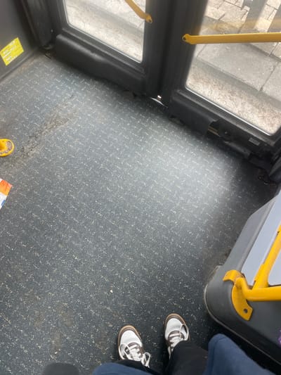 The scene depicts the interior floor of a public bus in Raanana, Israel, viewed from an overhead perspective. The floor is covered with dark grey, speckled anti-slip material, showing visible wear, scuff marks, and a concentrated dirty patch towards the lower-left quadrant. A small piece of litter, an orange and white wrapper, rests on the floor in the lower-left.

At the bottom of the frame, the feet of an individual are visible, wearing white and brown athletic shoes with white laces and dark trousers.

The front or side bus doors are visible in the upper half of the image, featuring black frames and glass panes that reveal a concrete sidewalk or street surface outside. A yellow, curved grab handle with a circular base is partially visible on the floor near the left door frame. On the right, a grey panel extends into the frame, with a horizontal yellow grab handle attached. The rubber seal at the bottom of the right door appears partially dislodged or damaged. Yellow rectangular warning labels are affixed to the black frame of the left door.