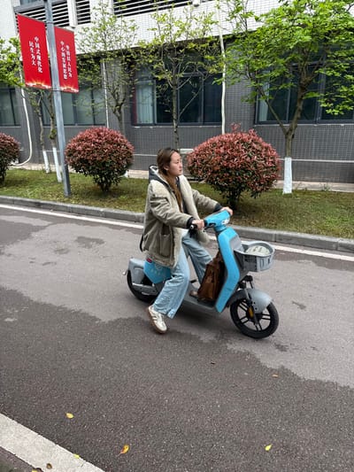 A young adult female is depicted riding a light blue electric scooter on an asphalt road in Jiangjin, China, on an overcast day. The individual, positioned slightly right of the frame's center, has light brown hair and is wearing an olive-green jacket with a cream-colored fleece lining, light blue baggy jeans, and light-colored sneakers with tan soles. Her hands are on the scooter's handlebars, and her gaze is directed towards the right. A brown leather satchel is suspended from the scooter's handlebars, and a light-colored object, possibly a cap, rests in the front basket. The asphalt road appears partially wet, with a white line visible on the left side and scattered yellow leaves. To the right of the road is a green median strip featuring rounded bushes with reddish-brown foliage and several deciduous trees with light green leaves, some with white-painted lower trunks. In the background, a multi-story building with a light gray facade and numerous dark-framed windows is visible. Two vertical red banners with yellow Chinese characters are attached to a pole behind the rider; the left banner reads "中国式现代化 民生为大" (Chinese modernization, people's livelihood is paramount), and the right banner reads "坚持以人民为中心的发展思想" (Adhere to the people-centered development philosophy).