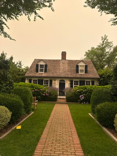 A Dutch colonial style house with cedar shake siding and a brick chimney is centered in the frame, with a brick pathway leading to its dark colored front door. The house has dark shutters on its windows, with dormer windows on the upper level. Lush green bushes and flowering plants, including hydrangeas, surround the house, with manicured lawns on either side of the pathway. Trees with dense foliage frame the top of the image. The sky is overcast with a hazy, orange hue, suggesting a smoky or dusty atmosphere. A small yellow marker is visible in the grass on the left side of the pathway. The address "82" is visible next to the front door. There are no people visible in the image. The location appears to be a residential area in Provincetown, Massachusetts, USA.