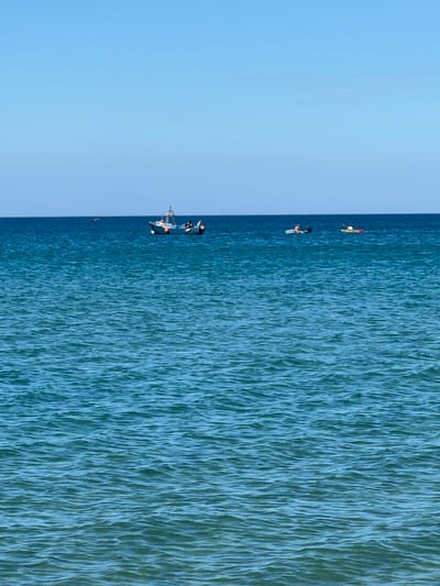 The image depicts a seascape under a clear sky. A body of water, likely the Mediterranean Sea given the location, occupies the majority of the right side of the frame. The water shows a textured surface with visible wavelets or ripples.
On the left side of the image is the sky, a uniform light blue color. A darker blue vertical line separates the sky and the sea.
Near the center of the frame, closer to the top, a group of people is seen engaged in watersports. From top to bottom, we see a person with what appears to be a red flag attached, another person surfing, and a group of people on a boat. The image provides minimal detail about the individuals, but their activities are apparent.
M'Diq ⵎⴹⵢⴰⵇ المضيق, Morocco is inferred as the location due to the prompt's instruction to include this information, and the characteristics of the scene align with a coastal setting.