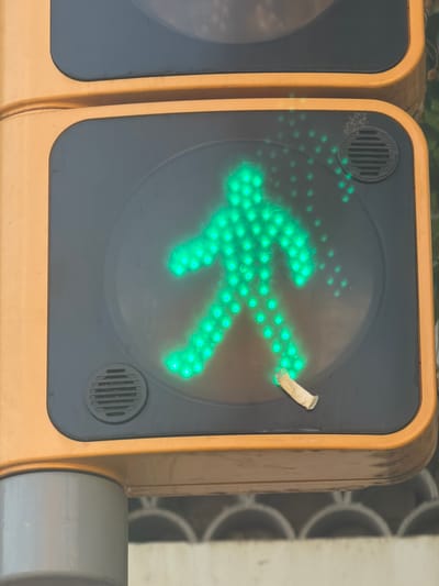 The image shows a close-up of a pedestrian traffic signal displaying a green walking figure. The signal is housed in a yellow casing, and a portion of an adjacent signal unit is visible to the left. The walking figure is composed of numerous small green lights, forming the silhouette of a person with arms and legs in motion. There is a small piece of white tape attached to the right side of the signal's display, partially obscuring some of the green lights. Two circular speaker grilles are present on the black display panel, one near the top left and another near the bottom right. The background is blurred, but appears to be an outdoor setting with some metal structures or fencing. The overall impression is that of a typical urban streetscape, likely in Barcelona, Spain, given the context. The lighting suggests it is daytime, and there are no obvious weather cues. The image focuses on the traffic signal itself, with no people visible. The presence of the tape might suggest a minor repair or temporary marking.