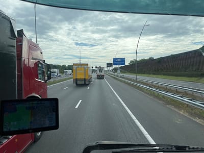 The image shows a view from inside a truck cab, looking out at a highway. In the foreground, on the left, is the red cab of the truck. To the right of the cab is a GPS device mounted on the windshield, displaying a map. The highway stretches ahead, with several trucks visible. A yellow truck with "JUMBO" written on it is in the middle lane. Another truck with a white trailer is in the right lane. A blue sign on the right side of the highway reads "Leenderheide 1200 m". The sky is overcast with clouds. The scene appears to be on a highway in or near Eindhoven, Netherlands, indicated by the sign for Leenderheide, a locality south of Eindhoven.