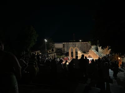 In Nesebar, Bulgaria, at night, a crowd of people is gathered in an outdoor amphitheater. The audience is seated on stone steps, facing a stage area. The stage features ancient Roman-style architecture with arches and brickwork, illuminated by spotlights. A performance appears to be underway, with several figures on stage wearing colorful costumes, potentially actors or performers. The overall atmosphere is one of a cultural event held at a historical site. The lighting is dim, with spotlights on the stage and some ambient light from streetlamps and nearby buildings. Trees are visible on the sides, casting shadows. The image captures a moment of communal gathering and entertainment within an ancient setting.