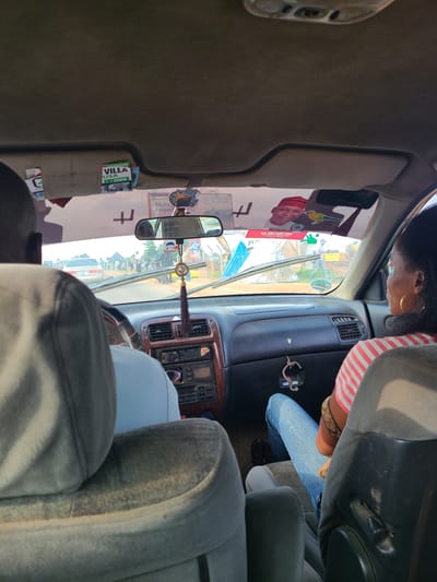 The image, taken from inside a car, shows the view through the windshield. The interior of the car has a beige headliner and a gray dashboard. A car stereo is visible in the center of the dashboard. A decorative tassel hangs from the rearview mirror. Two people are visible in the car: the driver, whose back is to the camera, and a woman in the passenger seat. Outside the car, a street scene is visible with other vehicles and people. Several signs are attached to the interior of the windshield. One sign has a picture of a man in a red hat and the text "ALI AHMED. KARRA YOUSUF." Another sign reads "VILLA USA ECFA." A third sign says "SAHAD HOSPITALS." The presence of these signs and the general street scene suggest the location is likely a busy area in Abuja, Nigeria, possibly a commercial or transportation hub.