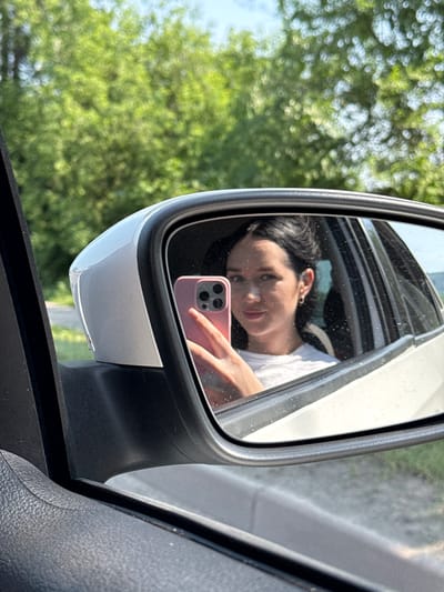 The image shows a young woman taking a selfie in a car's side mirror. She is inside the car, and her reflection is visible in the mirror. She is holding a pink smartphone up to take the photo. She has dark hair and is wearing a white t-shirt. The car appears to be parked or driving on a road surrounded by green trees and a bright sky. The location is not specified in the image.