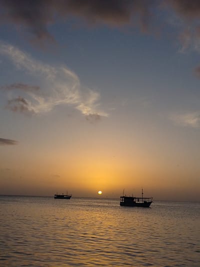 The image depicts a serene sunset scene over the waters of Juan Griego, Venezuela. The sky transitions from a deep blue at the top to a soft orange and yellow gradient near the horizon, illuminated by the setting sun. Wispy clouds, tinged with pink and gray, are scattered across the upper portion of the sky.
Two fishing boats are visible in the water. The boat on the right is larger and closer to the viewer, silhouetted against the vibrant sunset. It appears to be a traditional fishing vessel with a cabin structure and masts. The boat on the left is smaller and farther away, also silhouetted. The water is calm, with gentle ripples reflecting the warm hues of the sky. The overall impression is one of tranquility and natural beauty at the end of the day in this coastal Venezuelan location.