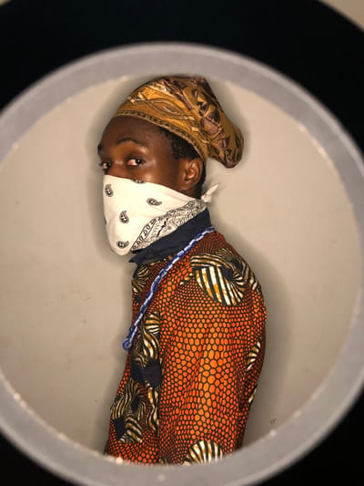 A person inside a circular frame wears a patterned orange and blue shirt. A white paisley bandana covers part of the face. The individual also wears a gold and brown head covering. A blue and white beaded necklace drapes across the chest. The person's eyes are visible above the bandana. The background appears to be a gray surface. The circular frame seems to be illuminated from the inside.