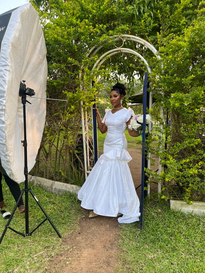 The image depicts a young Nigerian woman in a white wedding dress posing outdoors in Bukuru, Nigeria. She is standing under a decorative archway, holding a bouquet of white roses. The setting appears to be a garden or park, with lush green foliage surrounding the arch. A large white photographic softbox is visible in the foreground, suggesting that a photoshoot is in progress. The woman is looking downwards with a serene expression. The overall mood is calm and celebratory. The time of day appears to be daytime, with ample natural light. The weather seems pleasant. There is no visible text in the image.