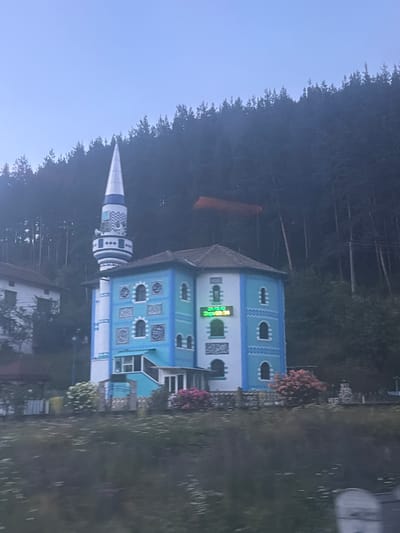 The image shows a mosque in Cherna Mesta, Bulgaria. The mosque is a light blue and white building with a tall, conical minaret. There is a digital clock on the side of the building that displays the time in green digits. The mosque is surrounded by trees, and the sky is a clear blue. The image appears to be taken from a moving vehicle, as the foreground is blurred.