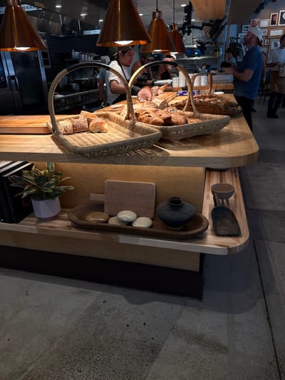 The image shows a bakery setup, likely a counter, with various food items displayed in baskets and on wooden boards. Several individuals are visible, including a man in a blue shirt near the counter and other people in the background, possibly employees or customers. The setting appears to be inside a restaurant or bakery.
Specifically, the image depicts goods sold at The Swedish Hill Bakery in Austin, Texas, USA.