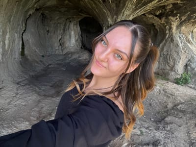 A woman stands in Skravena, Bulgaria, with her back to a cave. She wears a black long-sleeved shirt. Her brown and blonde hair is partially tied back. The cave walls are grey and rough.
