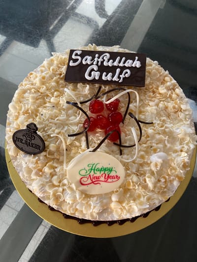 A round celebratory cake is centrally positioned on a golden, corrugated cardboard base, resting on a reflective, dark gray table surface. The cake's upper surface is densely covered with numerous small, irregularly shaped, light-colored flakes, consistent with white chocolate shavings or almond slivers. Dark chocolate frosting or drips are visible along the bottom circumference of the cake where it meets the golden board.

Key decorations include:
1.  A rectangular dark chocolate plaque positioned at the upper-center, displaying the text "Saifullah Gulf" in white icing.
2.  A cluster of approximately seven glossy red glacé cherries or maraschino cherries near the cake's center, surrounded by thin, curled strands of dark and white chocolate.
3.  A small, oval-shaped dark chocolate plaque located on the left side, embossed with "MR. BAKER" and a stylized chef's hat logo.
4.  A larger, oval-shaped white chocolate plaque situated at the lower-center, featuring "Happy New Year" written in decorative green and red script.

The reflective table surface shows indistinct, shadowed reflections. No individuals are visible, and no specific actions or interactions are depicted. The cake is branded by "MR. BAKER," a bakery chain with locations in Dhaka, Bangladesh. However, the image itself provides no visual information to identify a specific location within the city of Dhaka.