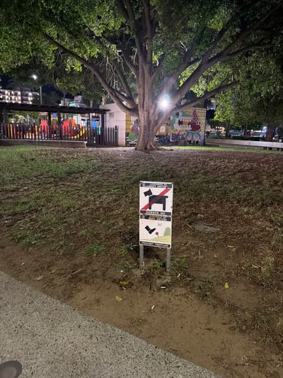 An outdoor park scene at night is captured. A large tree dominates the foreground, its branches extending over a grassy area. To the right of the tree, a two-part sign is mounted on a metal post. The top portion of the sign features a red circle with a diagonal line through a silhouette of a dog and a bench, indicating dogs are not allowed on benches. Text above reads "AJUNTAMENT DE SANT ANTONI". The bottom portion of the sign shows a red circle with a diagonal line through a footprint, indicating no walking on the grass. Text below reads "GOSSOS NO", "AJUNTAMENT DE SANT ANTONI", "Ai!", and "PROHIBIT TREPITJAR".

In the background, a playground with colorful structures is visible, enclosed by a wooden fence. Buildings and lights can be seen further in the distance. The ground is covered with grass and dirt, with fallen leaves scattered around. A paved path runs along the bottom left of the image. The lighting suggests artificial illumination from nearby sources.